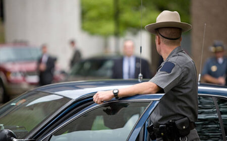 "A state trooper surveys a security detail while detectives, federal agents, and secret service mill about in the background"